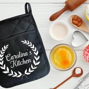 May include: Black quilted oven mitt with a white design of a wreath with the text "Carolina's Kitchen".