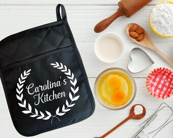 May include: Black quilted oven mitt with a white design of a wreath with the text "Carolina's Kitchen".