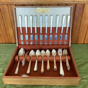 May include: A wooden storage chest containing a silver-plated flatware set. The interior features a cream-colored top section with a gold label reading Sheffield Silverplate Flatware and a rust-colored velvet base with slots for knives, spoons, and forks.
