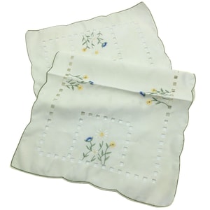 May include: Two off-white square napkins with scalloped edges and green trim. Each napkin features a central embroidered floral design with yellow, blue, and white flowers. The napkins have a cut-out square pattern around the floral design.