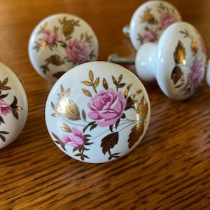 May include: A collection of white ceramic cabinet knobs with pink rose and gold leaf designs. The knobs have a rounded shape and are arranged on a wooden surface. The floral pattern adds a vintage touch.