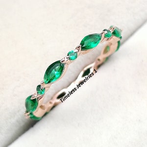 Puede incluir: Un delicado anillo de banda de oro rosa con una fila de piedras preciosas de color verde esmeralda en forma de marquesa. El anillo está engastado en un estilo simple y elegante.