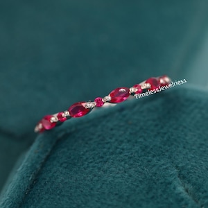 Könnte beinhalten: Ein zarter Silberring mit einer Reihe kleiner, ovaler Rubin-Edelsteine. Der Ring liegt auf einem grünen Samttuch.