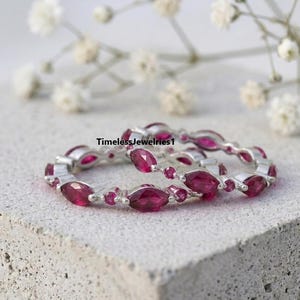 May include: Two silver eternity bands featuring alternating marquise-cut and round-cut deep pink gemstones. The rings are positioned on a textured stone surface with the text TimelessJewelries1 visible in the center of the frame.