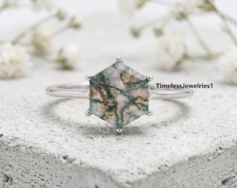 May include: A silver-toned ring featuring a hexagonal moss agate gemstone with intricate green and brown dendritic inclusions. The stone is held by six prongs on a thin, polished metal band. The text TimelessJewelries1 is visible on the right.
