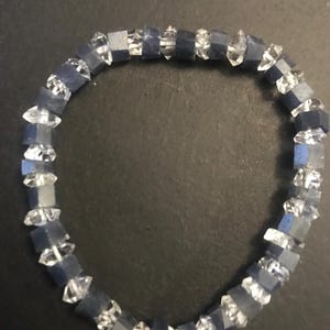 May include: A bracelet featuring alternating dark blue, square-cut beads and clear, faceted crystal beads. The bracelet is circular and rests on a dark surface, showcasing the contrast between the colors and textures of the beads.