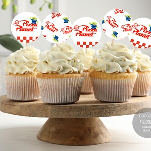 Puede incluir: Un conjunto de seis adornos para cupcakes con un diseño blanco, rojo y verde con las palabras "Pizza Planet" y un extraterrestre de dibujos animados. Los adornos están en un soporte para pasteles de madera.
