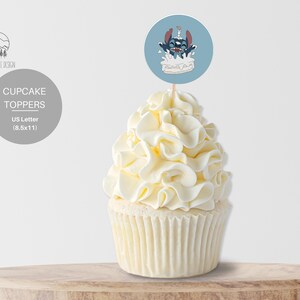 May include: Blue and white cupcake topper featuring a cartoon character with a blue body and red ears, holding a candle and sitting on a white cake. The topper is on a wooden surface.