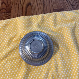 May include: A small, silver-colored metal bowl with a fluted design around the rim. The bowl is circular with a slightly raised center. The bowl is sitting on a yellow cloth with white polka dots.