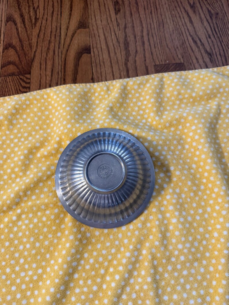 May include: A small, silver-colored metal bowl with a fluted design around the rim. The bowl is circular with a slightly raised center. The bowl is sitting on a yellow cloth with white polka dots.