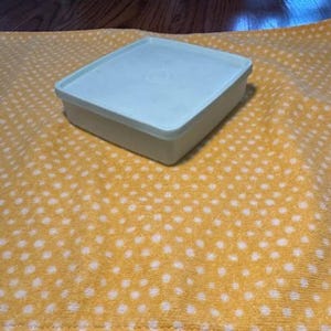 May include: A light blue square container with a matching lid. The container is sitting on a yellow towel with white polka dots. The background is a wooden surface.