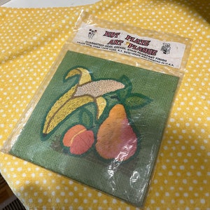 May include: A green hot plate plaque with a banana, pear, and flower design. The plaque is made of a heat, grease, and stain resistant material. The text on the packaging reads "Hot Plate Plaque" and "Guaranteed Heat, Grease, Stain Resistant Finish".