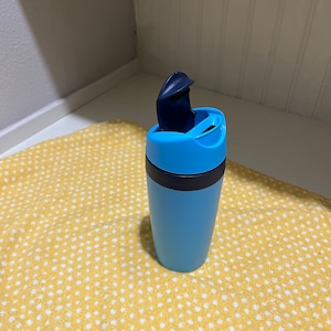 May include: A blue travel mug with a black lid and a blue flip top. The mug is sitting on a yellow and white polka dot surface.