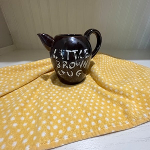 Pode incluir: Uma jarra de cerâmica marrom com letras brancas que dizem "Little Brown Jug". A jarra está sentada em um tecido amarelo com bolinhas brancas.
