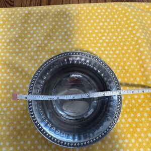 May include: A silver metal bowl with a beaded rim, approximately 6 inches in diameter, is measured with a tape measure. The bowl sits on a yellow fabric with white polka dots. The bowl has a reflective surface.
