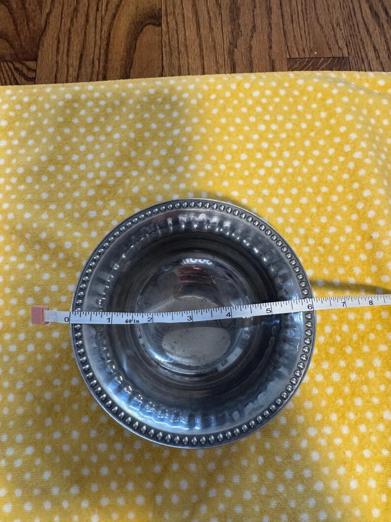May include: A silver metal bowl with a beaded rim, approximately 6 inches in diameter, is measured with a tape measure. The bowl sits on a yellow fabric with white polka dots. The bowl has a reflective surface.