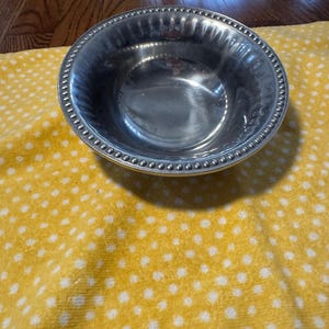 May include: A shiny, silver-colored bowl with a beaded rim. The bowl is round and has a slightly textured interior. It sits on a yellow cloth with white polka dots.
