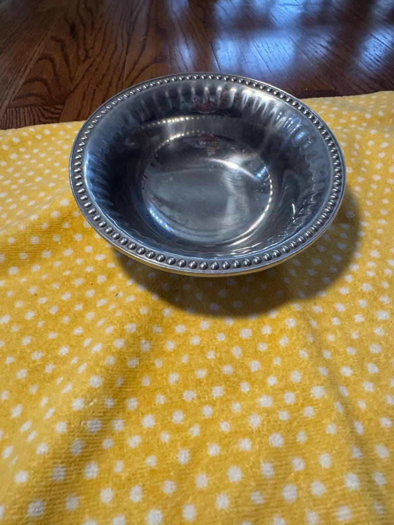 May include: A shiny, silver-colored bowl with a beaded rim. The bowl is round and has a slightly textured interior. It sits on a yellow cloth with white polka dots.