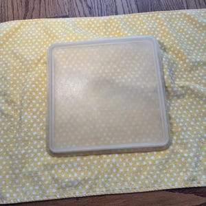 Puede incluir: Un recipiente de plástico transparente y cuadrado con tapa, sobre un paño amarillo con lunares blancos. El recipiente es translúcido y la tapa es ligeramente opaca. El fondo es una superficie de madera.