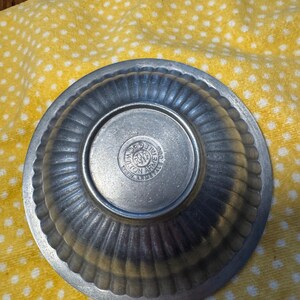 May include: A small, antique pewter bowl with a ribbed exterior and a circular base. The base features an embossed seal with text. The bowl is set against a yellow cloth with white polka dots. The bowl's design suggests it is vintage.