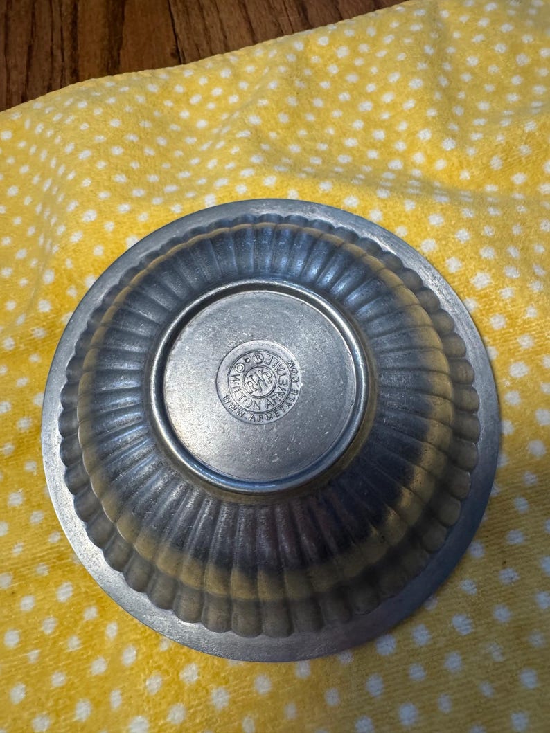 May include: A small, antique pewter bowl with a ribbed exterior and a circular base. The base features an embossed seal with text. The bowl is set against a yellow cloth with white polka dots. The bowl's design suggests it is vintage.