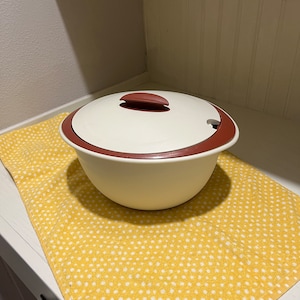Puede incluir: Un bol de servir de cerámica blanca con una tapa roja y un asa. El bol está sobre una toalla de cocina amarilla con lunares blancos.