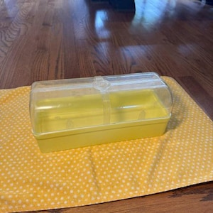 May include: A rectangular butter dish featuring a solid yellow opaque base and a clear, transparent plastic lid. The lid has a smooth, curved top with a subtle ribbed design centered on the middle, providing a clear view of the interior.