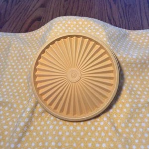 以下が含まれることがあります： 放射状のデザインが施された、丸い薄黄色のTupperwareの蓋。白い水玉模様の黄色い布の上に蓋が置かれています。背景は木製の表面です。