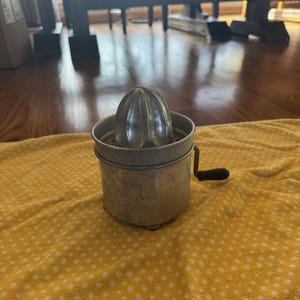May include: A vintage metal citrus juicer with a ribbed reamer and a small handle. The juicer sits on a yellow cloth with white polka dots. The juicer is designed to extract juice from citrus fruits.