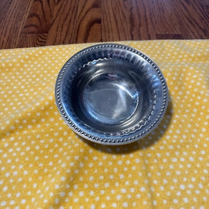 May include: A small, round, silver-colored bowl with a beaded rim. The bowl is sitting on a yellow cloth with white polka dots. The bowl is reflective and appears to be made of metal.