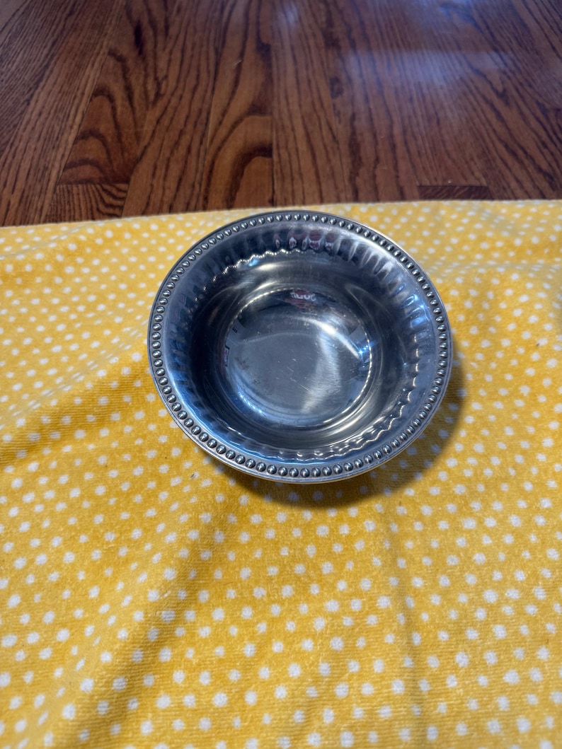 May include: A small, round, silver-colored bowl with a beaded rim. The bowl is sitting on a yellow cloth with white polka dots. The bowl is reflective and appears to be made of metal.
