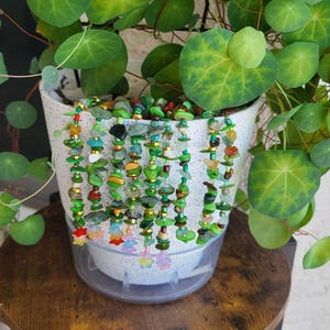 May include: A white speckled ceramic pot with a trailing plant and decorative beaded strands. The strands are made of various green, blue, and gold beads, with colorful flower-shaped charms at the bottom. The plant has round, green leaves.