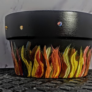 Puede incluir: Maceta de cerámica pintada de negro con un diseño de llama pintado a mano en tonos amarillos, naranjas y rojos. La maceta tiene gemas decorativas alrededor del borde. Una pieza única y artística para la decoración del hogar.