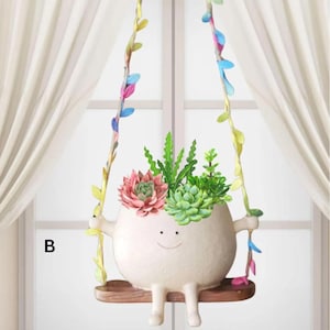 Op de afbeelding: Een grillige witte keramische plantenbak in de vorm van een lachend karakter, met een verscheidenheid aan vetplanten. De plantenbak hangt aan kleurrijke wijnstokken, opgehangen aan een houten schommel. De letter "B" is zichtbaar linksonder.
