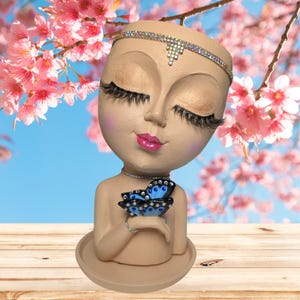 May include: A beige-colored decorative planter shaped like a woman's head and shoulders, with closed eyes and long eyelashes. The planter holds a blue butterfly and is adorned with a jeweled headband and necklace. The background features pink flowers.