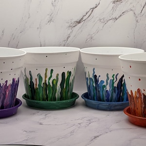 Puede incluir: Cuatro macetas de cerámica blanca con diseños pintados. Las macetas tienen una base blanca con un diseño colorido en la mitad inferior. Los diseños son morado, verde, azul y naranja. Cada maceta tiene un platillo a juego.