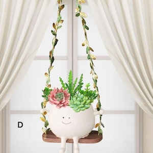 Op de afbeelding: Een grillige witte keramische plantenbak in de vorm van een lachend karakter, met een verscheidenheid aan vetplanten. De plantenbak hangt aan decoratieve wijnstokken met bladeren. De plantenbak staat op een kleine houten schommel.