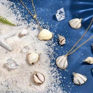 May include: A collection of gold-coloured seashell necklaces and pendants, set against a blue backdrop with sand and crystals. The necklaces showcase diverse shell designs, including cowrie and scallop shells, with gold chains.