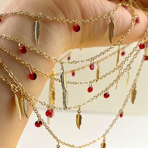 May include: Gold-toned layered necklace with red beads and feather charms. The necklace features multiple strands of delicate chain, each adorned with small, round red beads and detailed feather pendants. The jewelry is designed to be worn around the neck.