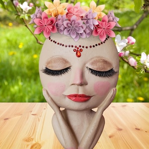 May include: A ceramic planter shaped like a woman's face with closed eyes and a delicate flower crown. The planter has a pink and white flower crown with red rhinestones around the forehead and a red rhinestone on the nose.