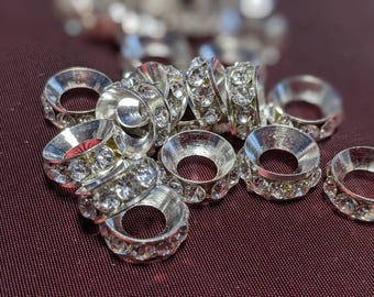 May include: Close-up of several silver-colored jewelry findings. Each finding has a circular design with a central hole and is encrusted with clear, sparkling rhinestones. The findings are scattered on a dark burgundy surface.