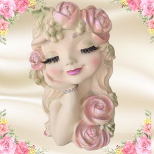 May include: A decorative, off-white face-shaped planter with closed eyes, long eyelashes, and a pink-painted mouth. The planter is adorned with pink roses and green foliage, and a rhinestone bracelet. The overall theme is floral and feminine.