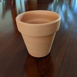 May include: A terracotta flower pot, unglazed, with a round opening and a slightly flared lip. The pot is empty and sits on a wooden surface.