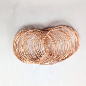 May include: A collection of multiple thin, circular bangles made of polished rose gold-colored metal. The bangles are stacked and overlapping, creating a dense, layered appearance with a reflective, metallic surface that catches the light.
