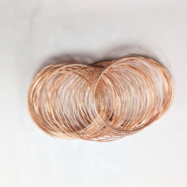 May include: A collection of multiple thin, circular bangles made of polished rose gold-colored metal. The bangles are stacked and overlapping, creating a dense, layered appearance with a reflective, metallic surface that catches the light.