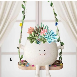May include: A whimsical white ceramic planter shaped like a smiling face, suspended on a wooden swing. The planter is filled with colorful faux succulents. The swing is decorated with green vines and small flowers.