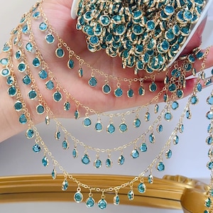 Op de afbeelding: Goudkleurige ketting met bungelende, teal-kleurige, ronde edelstenen. Er zijn verschillende strengen van de ketting zichtbaar, met een rol van dezelfde ketting op de achtergrond. De edelstenen zijn gelijkmatig verdeeld over de kettingen.