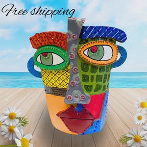 May include: A multi-colored ceramic face planter featuring a cubist-inspired design with vibrant orange, yellow, green, blue, and red sections. The surface is embellished with small rhinestones, a central gray nose strip, and red lips.