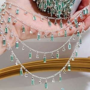 Könnte beinhalten: Silberne Ketten mit herabhängenden rechteckigen Smaragdgrün-Kristallen und kleinen klaren Kristallen. Der Schmuck wird in mehreren Strängen präsentiert, um das Design und den Glanz zu zeigen. Die Ketten werden in einer Hand gehalten, mit einem goldfarbenen Rahmen im Hintergrund.