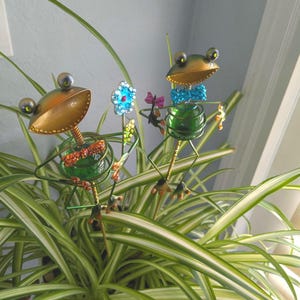 May include: Two decorative metal frog garden stakes featuring green glass bodies, gold-toned heads, and wire limbs. One frog wears an orange rhinestone bow tie, the other a blue rhinestone bow tie. A small metal flower with blue rhinestones stands between them.